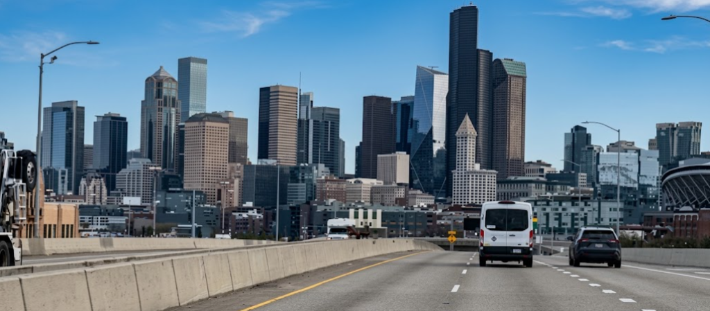 Eye-level perspective from a highway lane looking toward the downtown Seattle skyline, with a white van driving ahead on an elevated freeway against tall office towers and a blue sky.